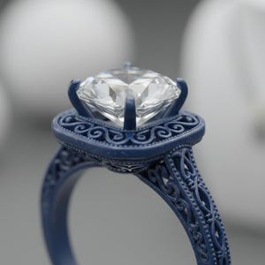 Puede incluir: Primer plano de un anillo de compromiso azul con un diamante grande y transparente. El anillo tiene un diseño de filigrana ornamentado en la banda y alrededor del engaste del diamante. El fondo está borroso.