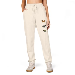 May include: Cream-colored sweatpants with a drawstring waist and three embroidered butterflies in shades of orange, black, and yellow. The sweatpants are a relaxed fit and have pockets.