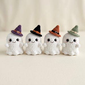 May include: Four small, white, crocheted ghost figures arranged in a row. Each ghost has large black safety eyes, a stitched smile, and pink blush marks. They wear miniature pointed hats in purple, black, orange, and sage green with small buckles.