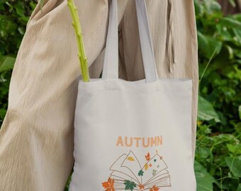 May include: A white tote bag featuring the word "AUTUMN" and the phrase "IN MY PAGES". The design includes an open book with autumn leaves. A yellow sunflower is placed inside the bag. The bag is held by a person.