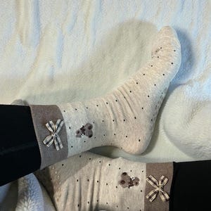 May include: A pair of light beige socks with small black polka dots. Each sock has a brown band at the top with a bow and a teddy bear applique. The socks are worn with black pants.