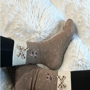 May include: A pair of brown socks with white polka dots, featuring a teddy bear applique and a small bow. The socks have a light beige cuff and are worn over black leggings, resting on a fluffy white surface.