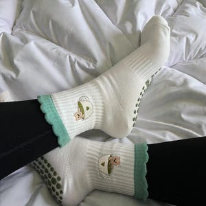 Könnte beinhalten: Weiße Socken mit hellgrüner, gewellter Bordüre und einem kleinen gestickten Design eines Teddybären in einer Teetasse. Die Socken haben grüne Anti-Rutsch-Griffe auf den Sohlen.