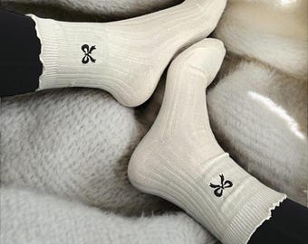May include: A pair of white cotton socks with a black bow embroidered on each sock. The socks have a ruffled edge.