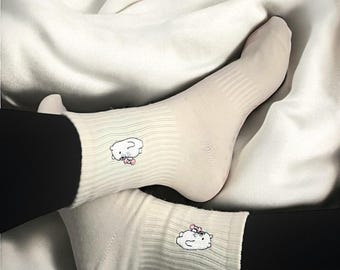 Puede incluir: Calcetines color crema con diseño acanalado, con una nube de dibujos animados con un lazo rosa. Se usan con leggings negros, sobre un fondo blanco texturizado. Un accesorio lindo y cómodo.