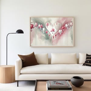 Puede incluir: Una pintura al agua enmarcada de flores de corazón sangrante en tonos de rosa y blanco, dispuestas en forma de corazón. La pintura está colgada en una pared blanca sobre un sofá blanco con almohadas marrones.