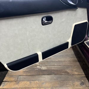 Puede incluir: Primer plano del interior de una puerta de coche con un panel de tela blanca y material de insonorización negro. El tirador de la puerta es visible y la puerta está abierta, revelando un suelo de madera.
