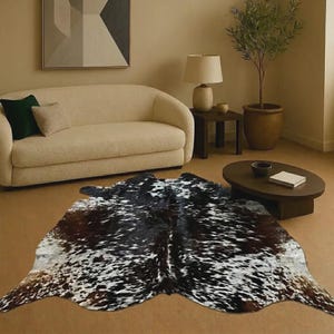 Puede incluir: Una alfombra de piel de vaca de forma irregular con un patrón moteado de pelaje negro, blanco y marrón. La alfombra está extendida sobre el suelo, mostrando una textura de piel animal natural con parches variados de colores oscuros y claros.