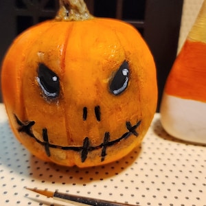 May include: A small orange pumpkin figurine with a hand-painted face featuring black triangular eyes with white highlights and a stitched mouth design. The pumpkin has a textured surface and a brown stem. A thin paintbrush lies in the foreground.