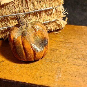 May include: A small, round, orange pumpkin figurine with a textured surface, visible vertical ridges, and a brown stem. The pumpkin features patches of dark brown and white coloring, giving it a weathered appearance with a realistic, matte finish.