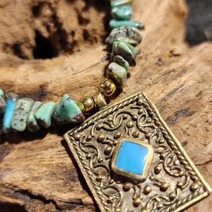May include: A necklace featuring turquoise-colored stone beads and a square pendant. The pendant has an ornate, gold-colored frame with a small, square, light blue stone in the center. The necklace is displayed on a piece of wood.