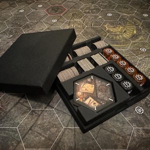 May include: A black foam insert for a board game box with compartments for cards, dice, and tokens. The insert is designed to organize and store game components.