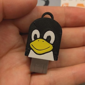 Könnte beinhalten: Ein schwarz-weißer USB-Stick in Form eines Pinguins mit gelbem Schnabel. Der Pinguin hat oben eine Schlaufe. Der USB-Anschluss ist grau.
