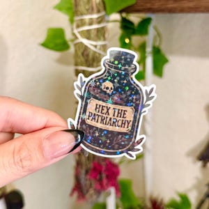 May include: A holographic sticker in the shape of a potion bottle. The bottle is black with a skull and star design. The sticker has the text "HEX THE PATRIARCHY" in a gold-colored font.