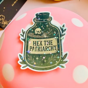 Puede incluir: Una pegatina que representa una botella de poci&oacute;n verde oscuro con una calavera y estrellas en su interior. La etiqueta dice "HEX THE PATRIARCHY" en letras doradas. La pegatina tiene un efecto hologr&aacute;fico y brillante, colocada sobre una superficie rosa.