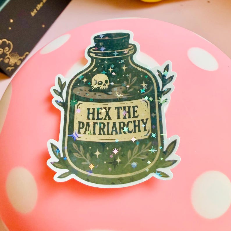 Puede incluir: Una pegatina que representa una botella de poci&oacute;n verde oscuro con una calavera y estrellas en su interior. La etiqueta dice "HEX THE PATRIARCHY" en letras doradas. La pegatina tiene un efecto hologr&aacute;fico y brillante, colocada sobre una superficie rosa.