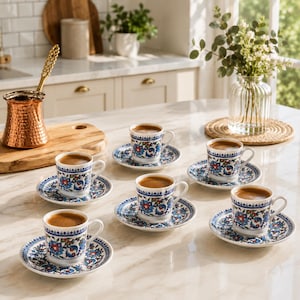 May include: A set of six white ceramic coffee cups and matching saucers featuring a blue, red, and green floral pattern, accompanied by a copper coffee pot with a textured finish, arranged on a light-colored marble countertop surface.