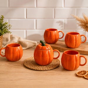 Peut inclure: Ensemble de tasses en céramique orange en forme de citrouille et un récipient en forme de citrouille avec couvercle. Les tasses ont une anse et sont disposées sur une surface en bois avec un dessous de plat tressé.