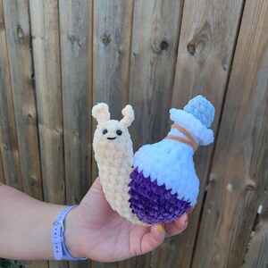 May include: A crocheted snail toy with a cream-colored body and a purple and white potion bottle shell. The snail has a smiling face and small antennae. The potion bottle is detailed with a brown string and a light blue cap.