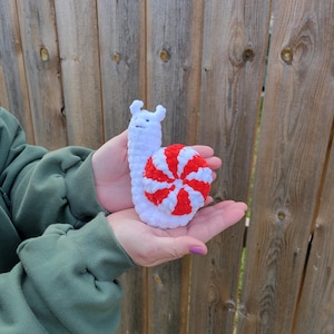 May include: A handmade crochet snail toy. The snail's body is white, and the shell is red and white striped. The snail has a smiling face and is held in open hands against a wooden fence background. The toy is soft and plush.