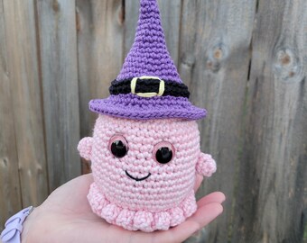 May include: A handmade pink crochet creature wearing a purple witch hat with a black band and gold buckle. The creature has large black eyes and a smiling mouth. The item is a Halloween decoration.