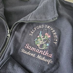 May include: Dark navy fleece zip-up jacket with embroidered details. Features "Salford University" text, a stethoscope, floral elements, and the name "Samantha, Student Midwife" in pink.