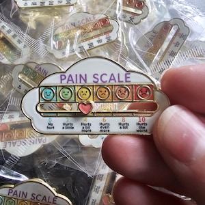 May include: A cloud-shaped enamel pin featuring a pain scale with six coloured faces ranging from blue to red. A sliding pink heart marker indicates pain levels from 0 to 10, with text labels including No hurt, Hurts a little, Hurts a bit more, and Hurts the worst.