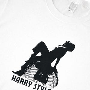 May include: A white short-sleeved t-shirt featuring a black graphic print of a person sitting on a disco ball. The text "HARRY STYLES" is printed in black below the graphic. The shirt has a crew neckline and a white fabric care label at the collar.