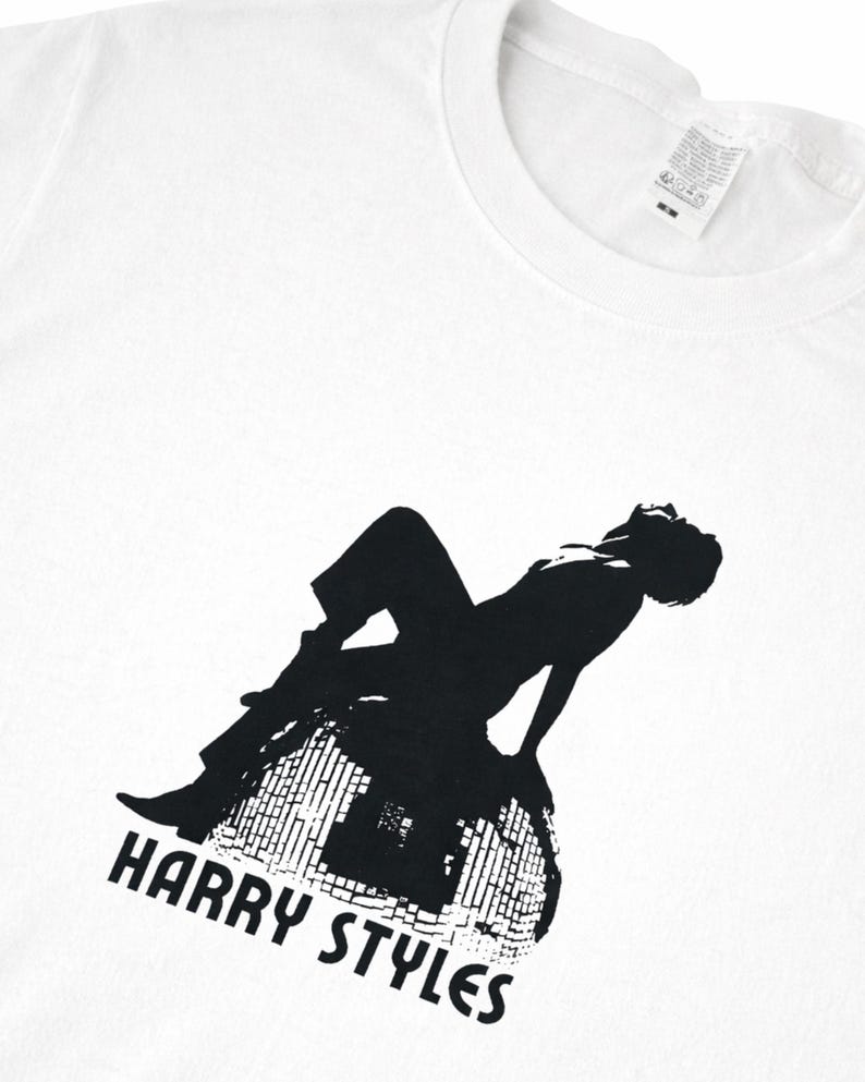 May include: A white short-sleeved t-shirt featuring a black graphic print of a person sitting on a disco ball. The text "HARRY STYLES" is printed in black below the graphic. The shirt has a crew neckline and a white fabric care label at the collar.