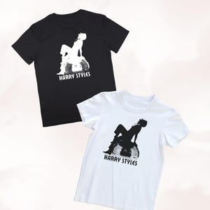 May include: A black t-shirt and a white t-shirt, each featuring a graphic print of a person sitting on a disco ball with the text HARRY STYLES printed underneath in a bold, sans-serif font. The black shirt has a white graphic, and the white shirt has a black graphic.
