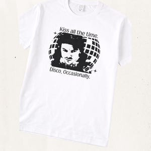 May include: A white short-sleeve t-shirt featuring a black graphic print of a person in front of a disco ball. The text above the graphic reads Kiss all the time. and the text below the graphic reads Disco, Occasionally. with small star icons.