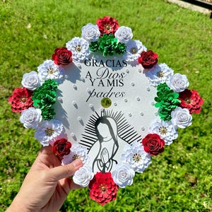 May include: A white graduation cap decorated with red, white, and green paper flowers. The cap features the text "GRACIAS A Dios Y A mis padres" and an illustration of a religious figure.