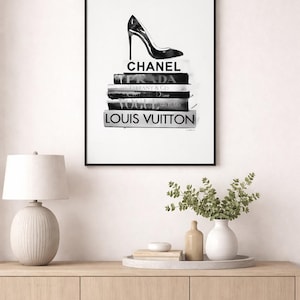 Puede incluir: Impresi&oacute;n en blanco y negro enmarcada que presenta un zapato de tac&oacute;n alto sobre una pila de libros de moda. Los libros muestran los nombres Chanel, Prada, Dior, Vogue y Louis Vuitton. La obra de arte se exhibe en una pared blanca.