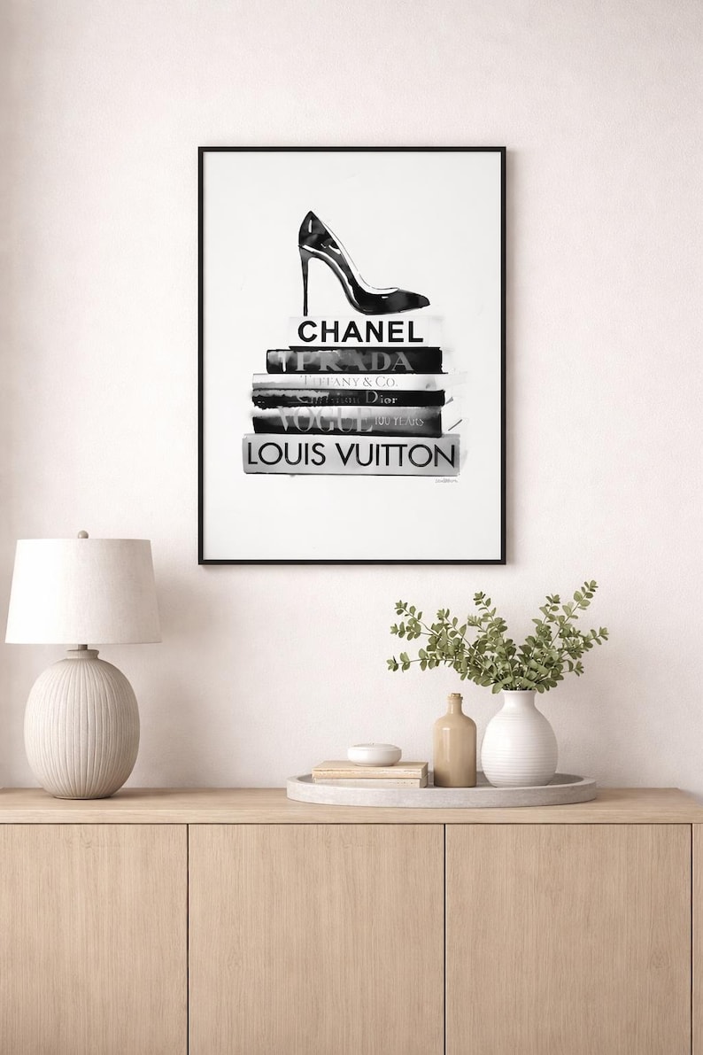 Puede incluir: Impresi&oacute;n en blanco y negro enmarcada que presenta un zapato de tac&oacute;n alto sobre una pila de libros de moda. Los libros muestran los nombres Chanel, Prada, Dior, Vogue y Louis Vuitton. La obra de arte se exhibe en una pared blanca.