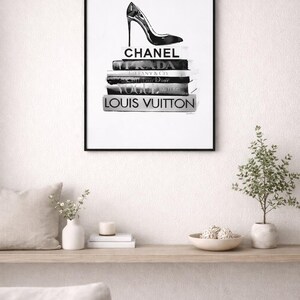 Puede incluir: Impresi&oacute;n en blanco y negro enmarcada que presenta un zapato de tac&oacute;n alto sobre una pila de libros de moda. Los libros est&aacute;n etiquetados con nombres de dise&ntilde;adores como Chanel, Prada y Louis Vuitton. La impresi&oacute;n se muestra en una pared blanca.