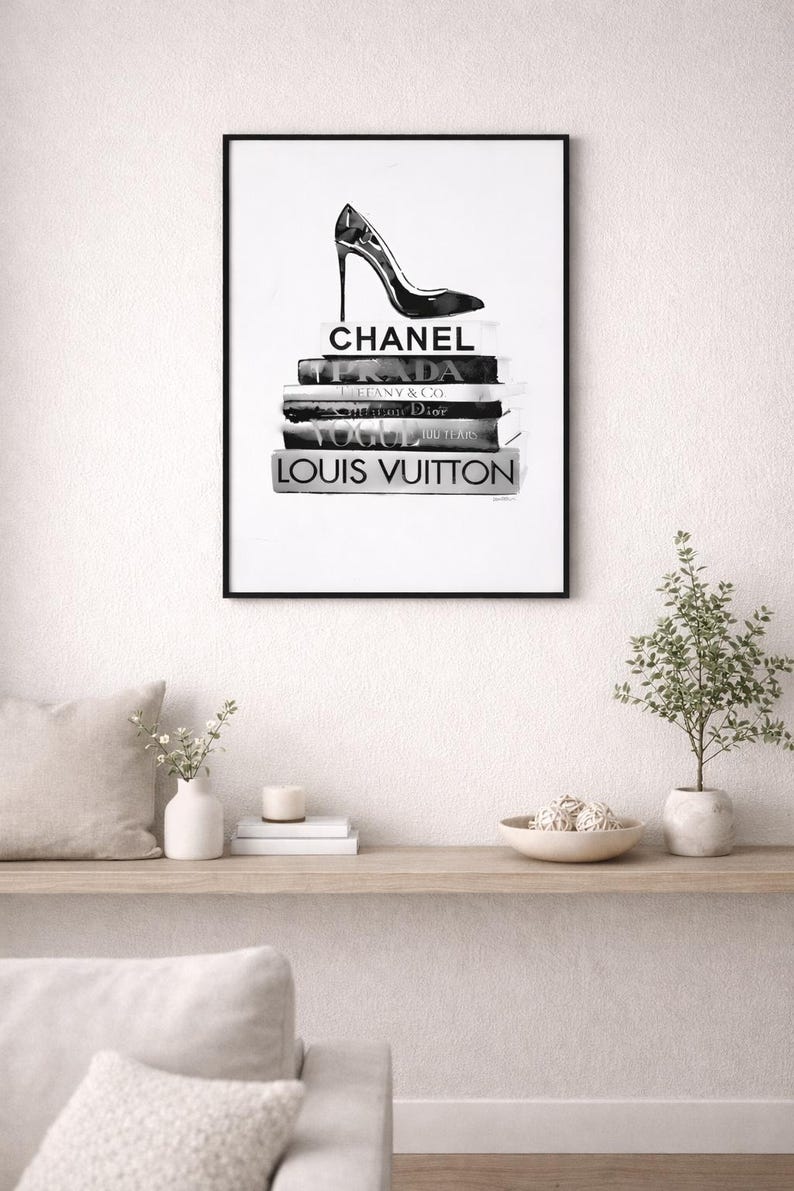 Puede incluir: Impresi&oacute;n en blanco y negro enmarcada que presenta un zapato de tac&oacute;n alto sobre una pila de libros de moda. Los libros est&aacute;n etiquetados con nombres de dise&ntilde;adores como Chanel, Prada y Louis Vuitton. La impresi&oacute;n se muestra en una pared blanca.