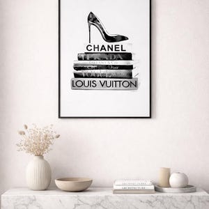 Puede incluir: Impresi&oacute;n en blanco y negro enmarcada que muestra un zapato de tac&oacute;n alto sobre una pila de libros de moda, incluyendo Chanel y Louis Vuitton. La impresi&oacute;n cuelga en una pared blanca sobre una superficie de m&aacute;rmol.