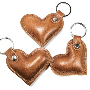 May include: Three brown leather heart-shaped keychains with silver key rings. The keychains have a textured surface and are stitched around the edges. The key rings are attached with a silver rivet.