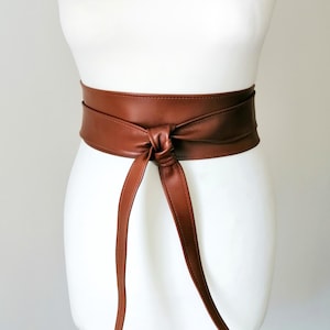 May include: A wide, smooth, brown faux leather obi-style belt wrapped around a mannequin. The belt features a central knot detail with two long, flat, tapered ends hanging down vertically from the front, creating a clean and structured look.
