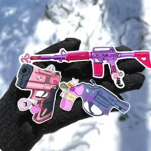 May include: A collection of stickers featuring stylized firearms. The stickers include a pink handgun, a purple and pink rifle, and a blue revolver. Each sticker has decorative elements and cartoon character accents.