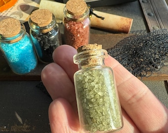 May include: Four small glass bottles filled with different colored crystals. The bottles have cork stoppers and are labeled with the words "WitchHippie Crafts" and "Ritual Salts". The labels also list the colors of the salts and their associated meanings.