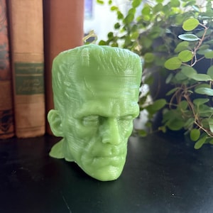 May include: A green candle shaped like Frankenstein's monster's head. The candle has a wick on top and is set against a backdrop of old books and greenery. The candle is a light green color.
