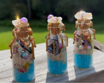 May include: Three decorative glass bottles with cork stoppers. Each bottle contains layers of blue liquid, sand, and various small objects. The bottles are adorned with decorative elements and a golden-colored wax. The bottles are displayed on a wooden surface.