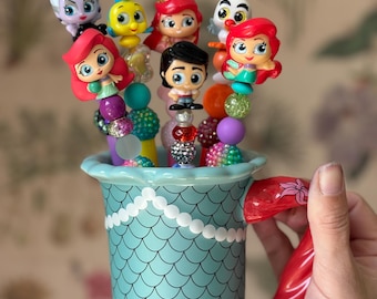 May include: A teal ceramic mug with a mermaid scale design, holding beaded pens topped with Disney characters. The mug has a red handle shaped like a mermaid tail. The pens feature characters like Ariel, Flounder, and Ursula.