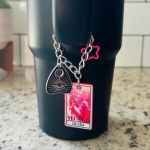 May include: A black travel mug with a silver chain attached to it. The chain has a pink star charm, a silver charm with an eye and a black and pink tarot card charm with the number XIII and the word "DEATH" on it.