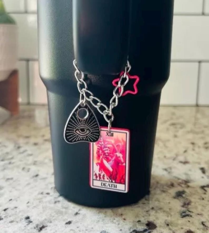 May include: A black travel mug with a silver chain attached to it. The chain has a pink star charm, a silver charm with an eye and a black and pink tarot card charm with the number XIII and the word "DEATH" on it.