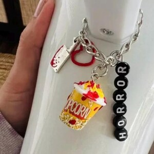 May include: A silver chain with charms, including a red heart, a silver knife, a popcorn bucket with red and yellow accents, and black beads that spell "HORROR".