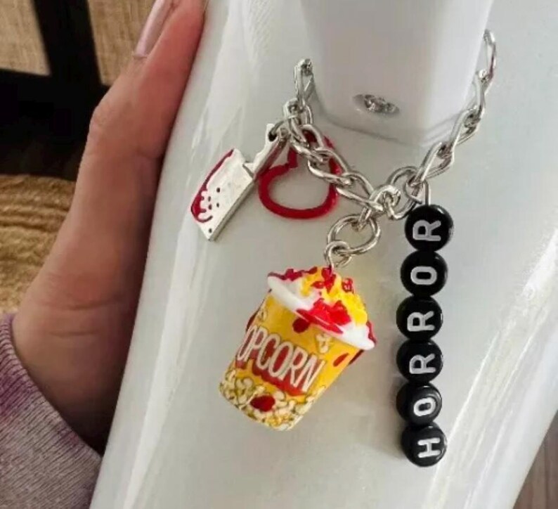 May include: A silver chain with charms, including a red heart, a silver knife, a popcorn bucket with red and yellow accents, and black beads that spell "HORROR".