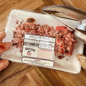 May include: A package of raw ground meat labeled "Mystery Meat" with a "Fresh Ground Daily" sticker. The package is on a wooden surface, with a pair of metal tongs and a snake skin in the background.