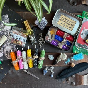 May include: A collection of mystical items including candles, crystals, a small sword, and a tin labeled "Mini Altar Kit." The candles are various colors, and the kit includes a variety of spiritual tools and accessories. The overall theme is related to witchcraft and spirituality.